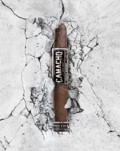 A Camacho Triple Maduro cigar rests on cracked concrete, showcasing its rich, dark wrapper and bold branding against a stark background.
