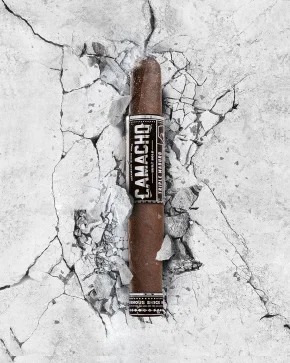 A Camacho Triple Maduro cigar rests on cracked concrete, showcasing its rich, dark wrapper and bold branding against a stark background.