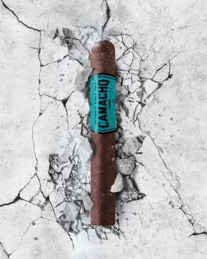 A Camacho Equador cigar emerges from cracked concrete, exuding a sense of strength and boldness with its distinctive orange band.