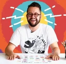 A man wearing glasses and a white t-shirt sits at a table surrounded by colorful stickers.