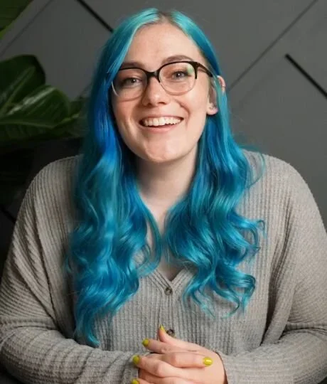 A woman with blue hair and glasses sits in front of a green plant, smiling softly at the camera.