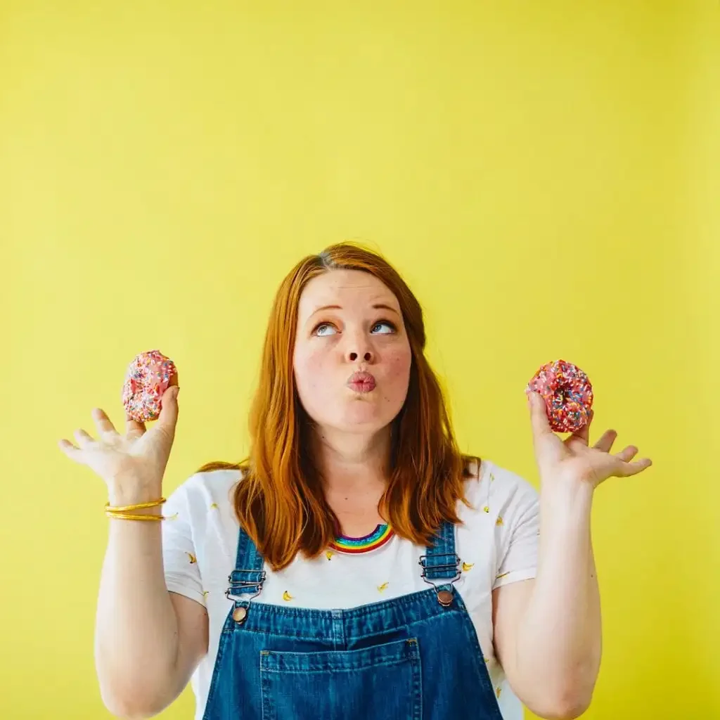 Playful Skillshare illustrator and teacher in overalls holding two sprinkled donuts against yellow background, inspiring creativity for beginners.