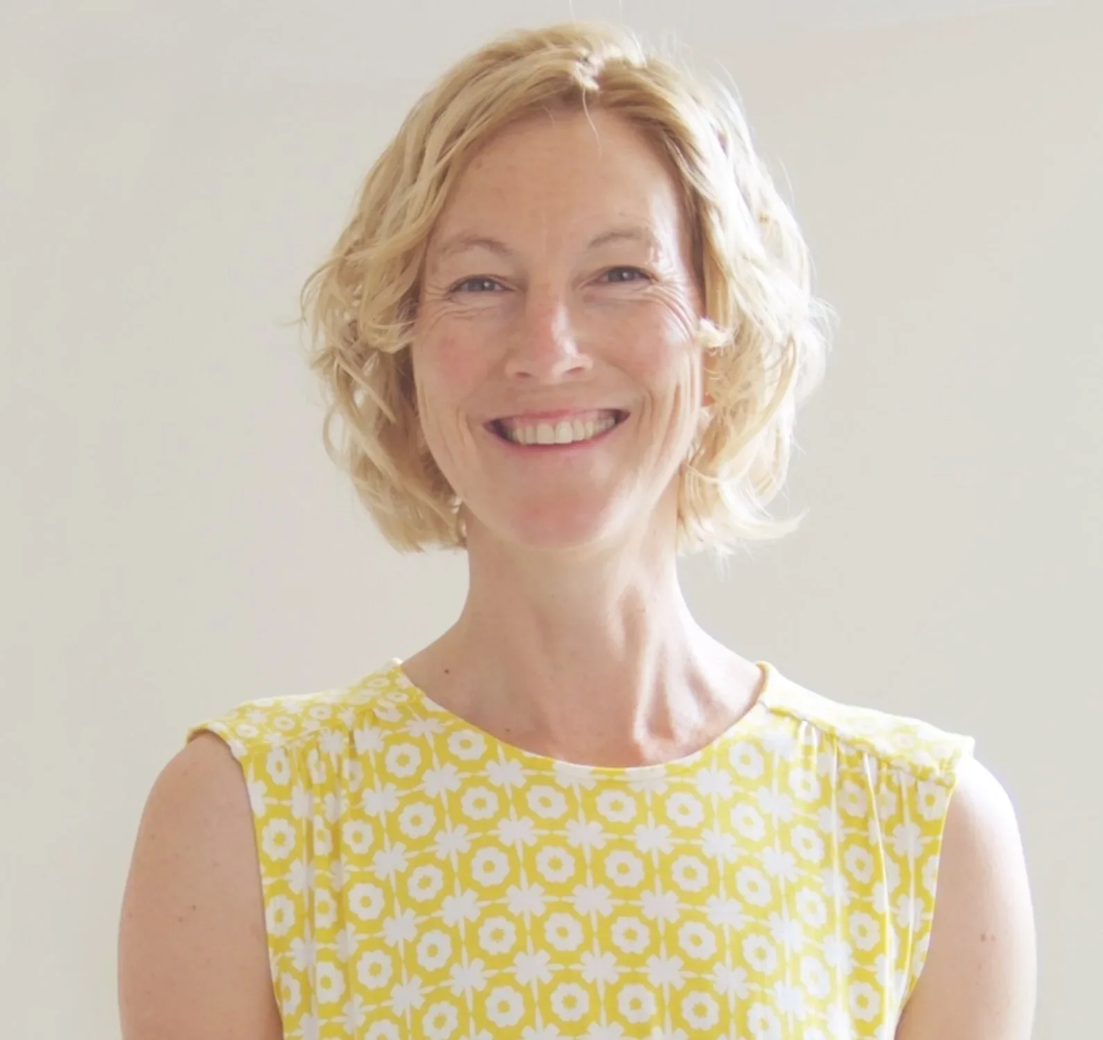 A woman with blonde hair in a yellow and white patterned dress stands smiling against a neutral background.