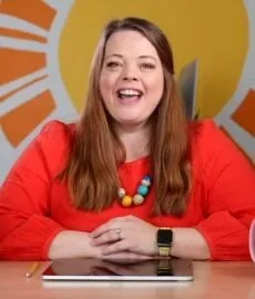An excited woman in a red shirt sits at a table, focused on a tablet in front of her.