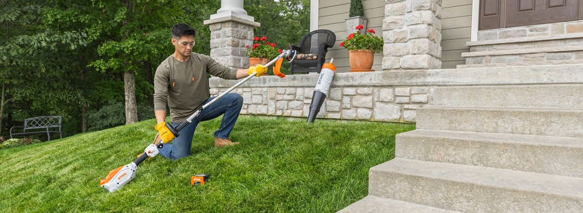 Man kneeling on grass with string trimmer and leaf blower