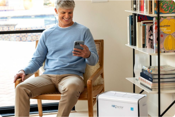 A middle-aged man with short gray hair, wearing a light blue sweater and beige pants, is sitting in a wooden chair indoors. He is smiling while looking at his smartphone. Next to him, on the floor, is an AirDoctor air purifier with a sleek white design. Behind him, there is a bookshelf filled with books and decorative items, and a large window showing an outdoor patio with a pool. The scene highlights a clean and comfortable living space with purified air.