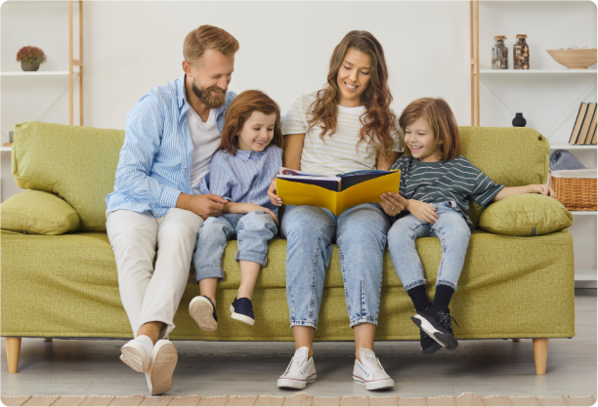  A happy family of four sits together on a green couch in a cozy living room. The mother holds an open book, reading aloud to her two young children, who are listening attentively with smiles. The father, sitting beside them, is also engaged in the moment. The scene reflects a clean, comfortable home environment, emphasizing the importance of fresh air and well-being—something AirDoctor air purifiers help provide. 