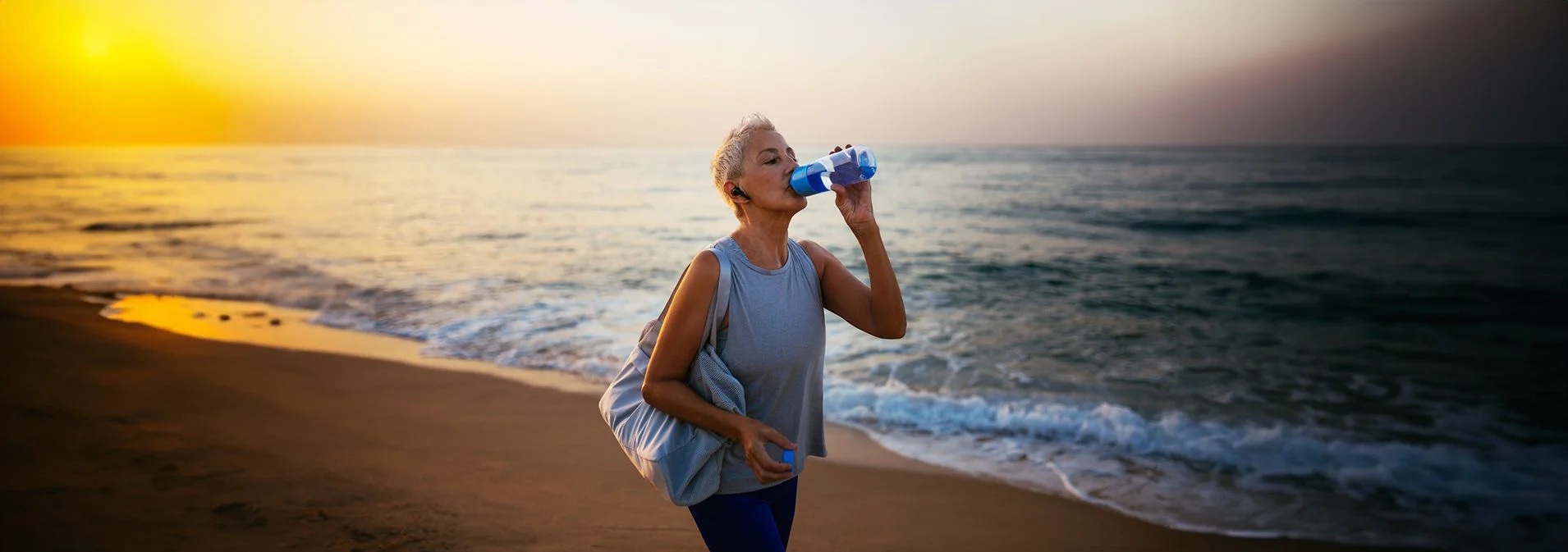 Older woman walking on the beach at sunset drinking out of a water bottle