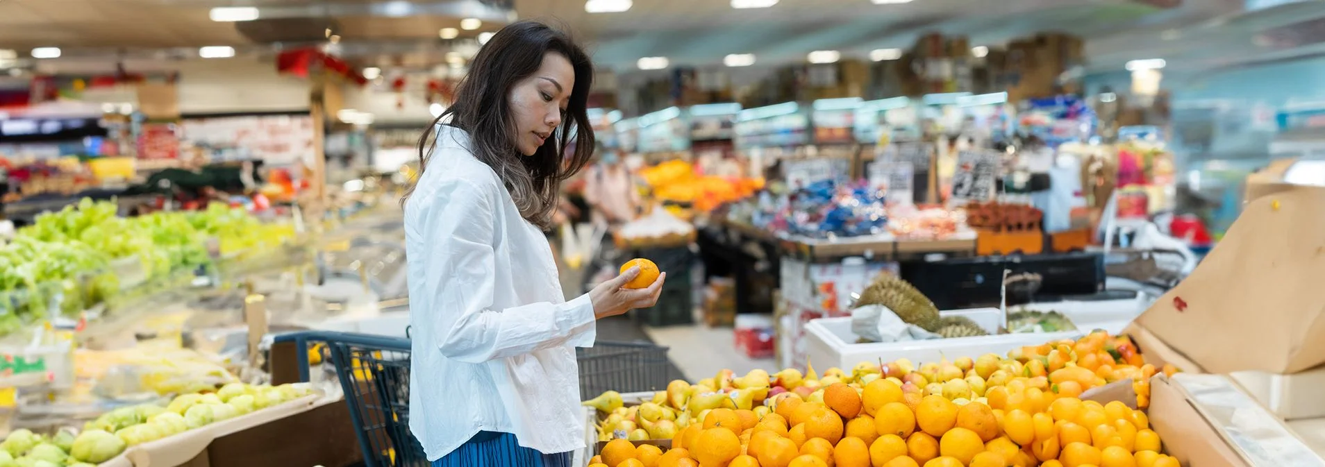A woman looking at an orange in the produce section of a grocery store