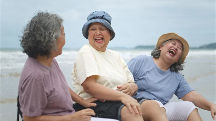 3 older women laughing while sitting on the beach