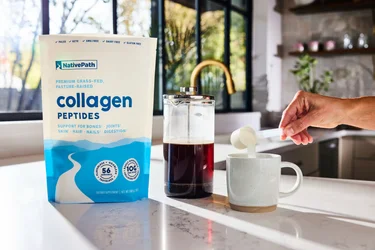 A hand pouring a scoop of NativePath Original Collagen Peptides from a large bag container into a coffee mug with a French Press in the background.