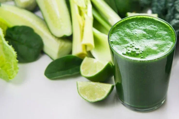 Clear glass of Leafy Greens Detox Smoothie with sliced cucumbers, limes, celery, and spinach in the background