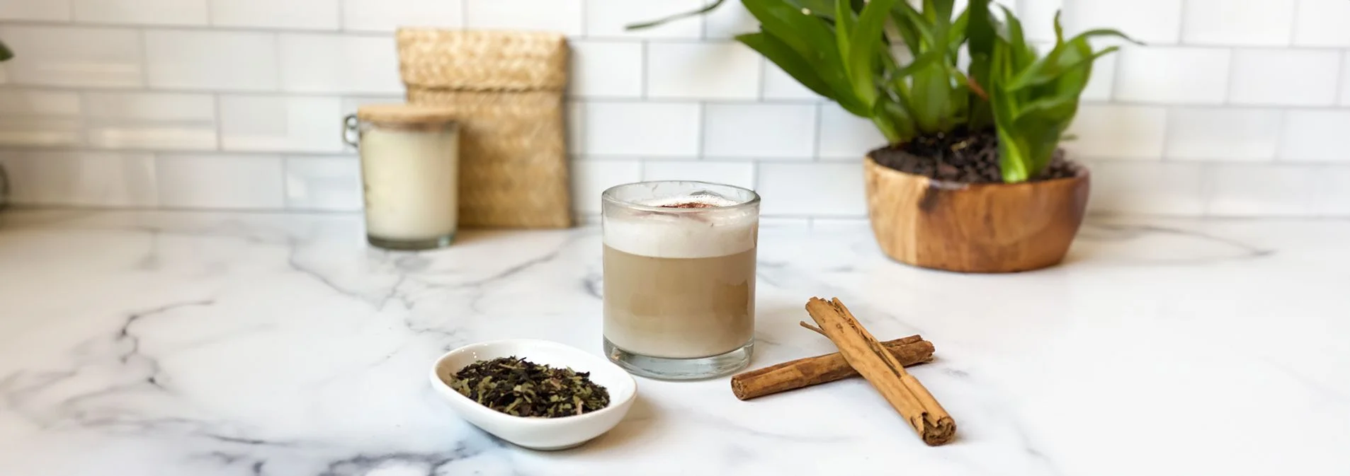 A front view of Vanilla Chai Latte in a clear glass mug with chair tea leaves in a bowl and cinnamon sticks next to it.