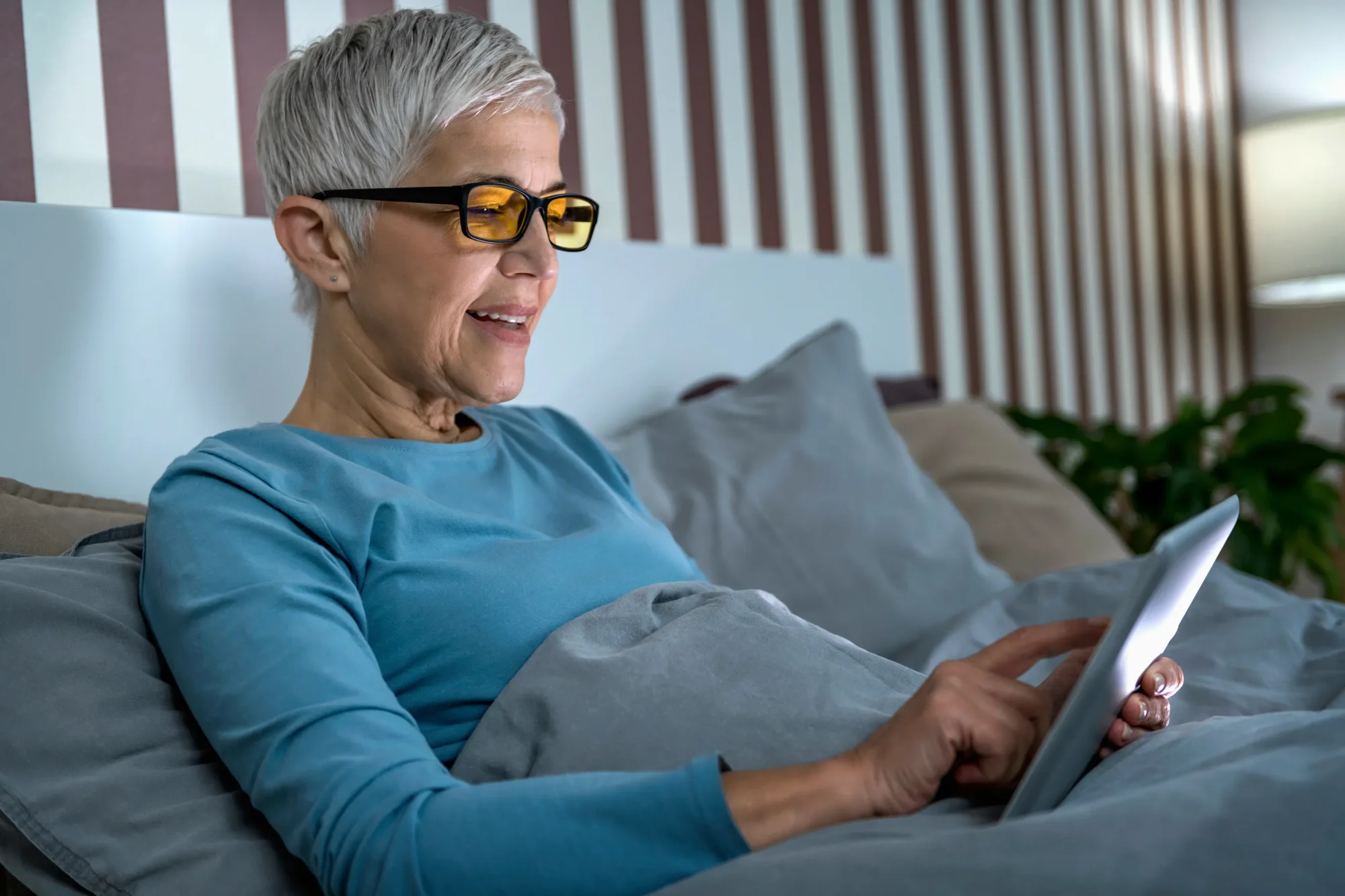 Older woman with short gray hair sitting in bed with a tablet and blue light-blocking glasses on.