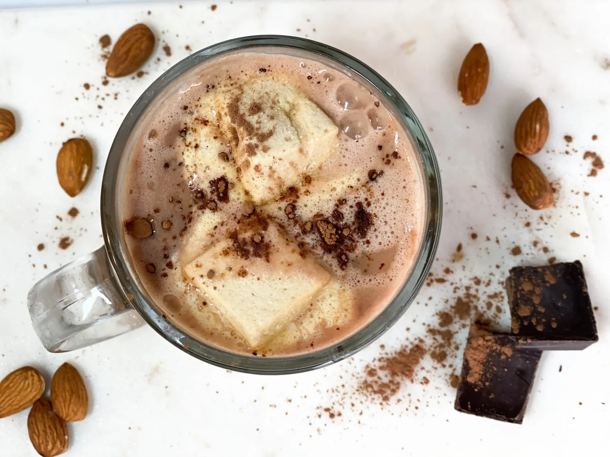 Top of view image of a mug of warm healthy hot chocolate with chocolate collagen. Marshmallows and a sprinkle of cacao are sprinkled on top. Almonds, cacao, and chunks of dark chocolate are sprinkled around the mug on a marble surface.
