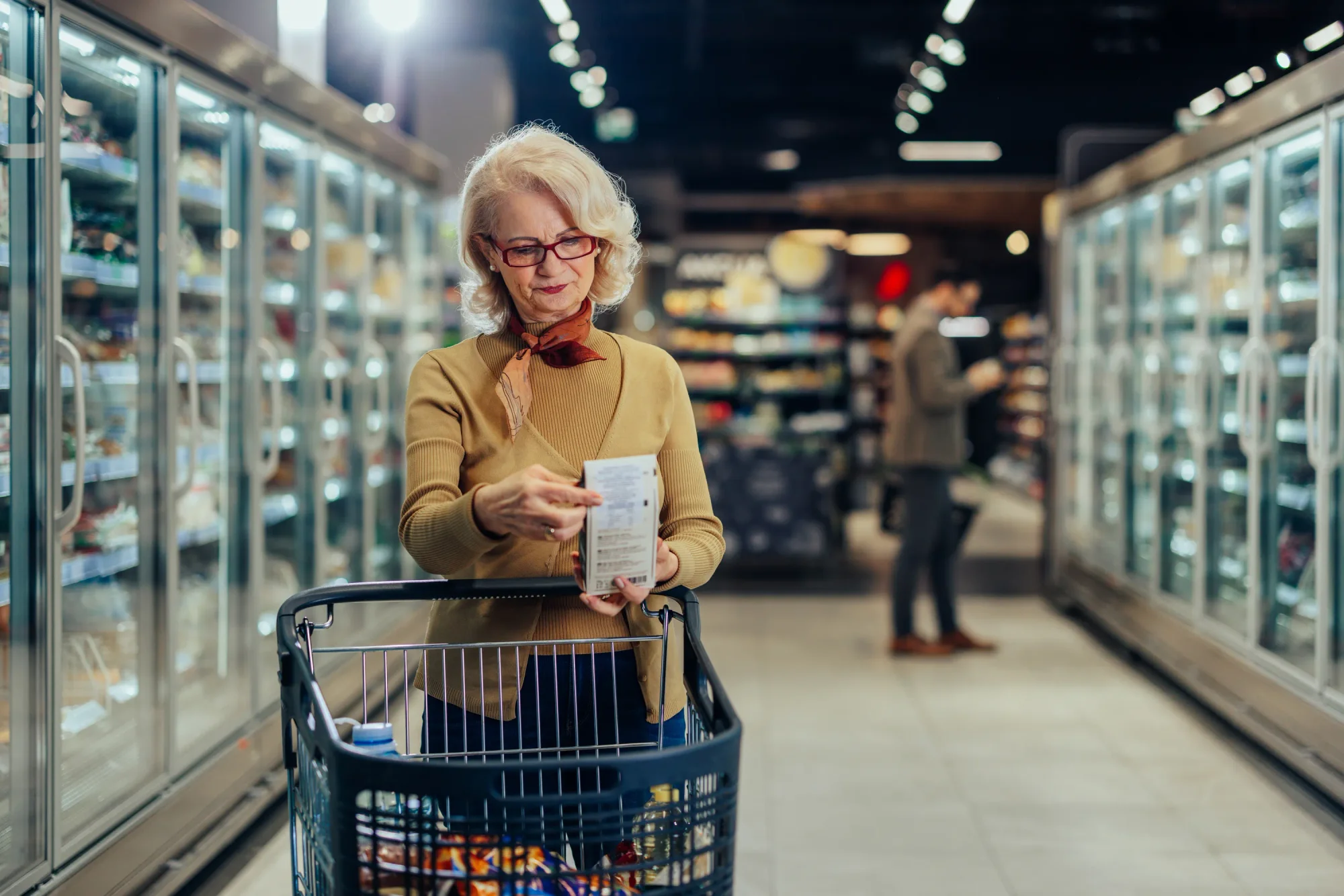 Senior woman shopping in a grocery store, standing beside shopping cart. She is reading the ingredient label on product.