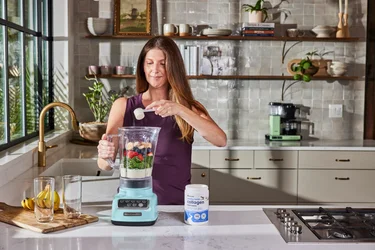 A woman pouring a scoop of NativePath Original Collagen Peptides into a blender full of fruits and vegetables