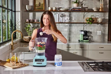 A woman pouring a scoop of NativePath collagen into a blender full of fruits and veggies