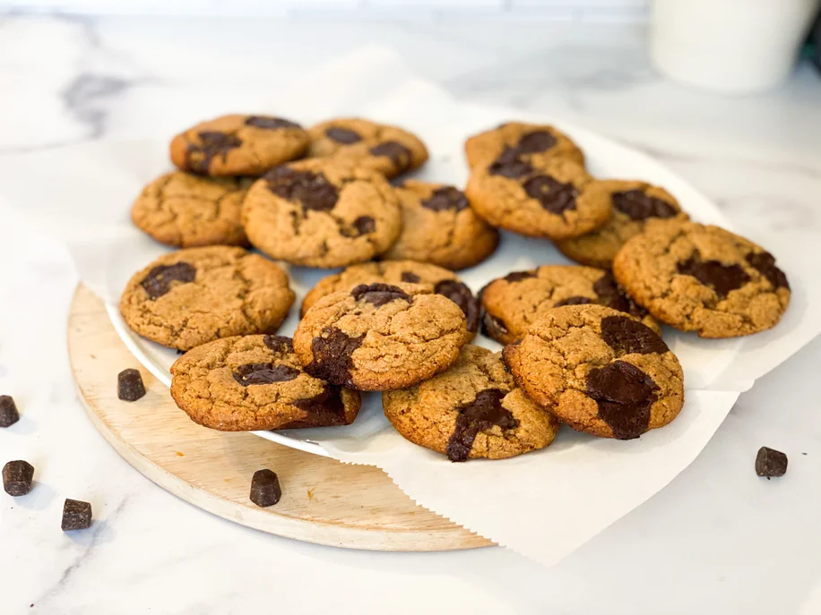 A plate of chocolate chip cookies on a wooden cutting board surrounded by chocolate chips.