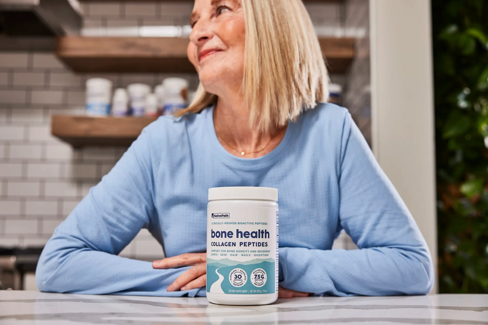 Older blonde woman in blue shirt sitting at granite kitchen countertop with jar of NativePath Bone Health Collagen Peptides.