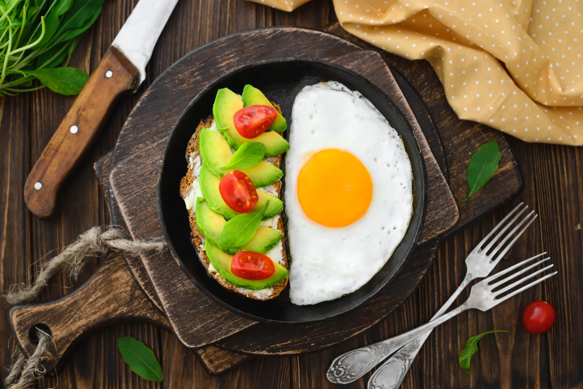 Fried egg in pan alongside avocado toast top view close-up in rustic style on a wooden background, cutting boards, forks, knife, arugula, concept of a healthy lifestyle with a high-fat, high-protein breakfast.