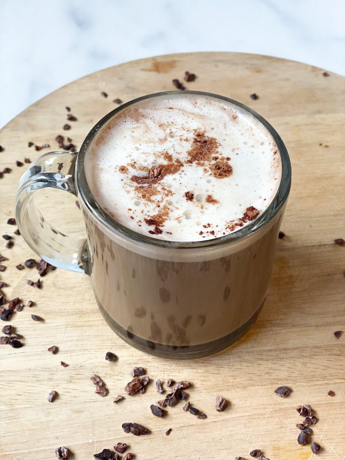 A close up view of the Mocha MCT Latte in a clear glass mug on a wooden board surrounded by coffee beans.