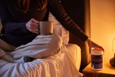 A woman in bed holding a mug and placing a container of NativePath Collagen PM on a nightstand.