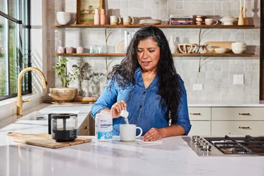 A woman pouring a scoop of NativePath Original Collagen Peptides into a blender full of fruits and vegetables