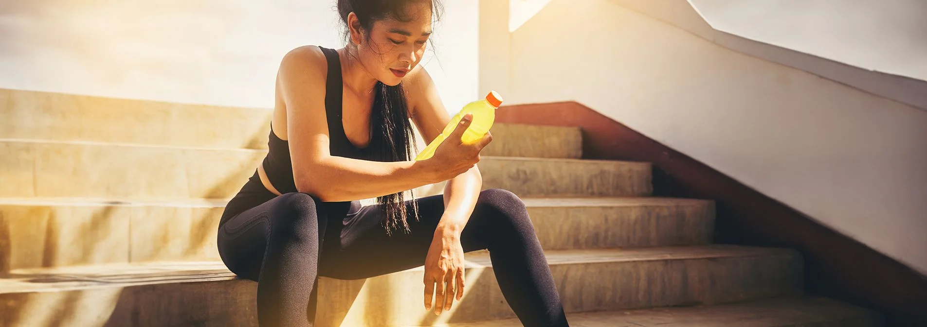 A woman sitting on the steps in workout clothes holding a bottle of water containing BCAA mix