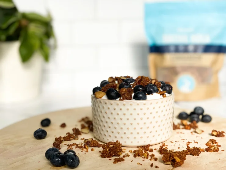 Bowl of plain dairy-free yogurt topped with Native Nola and fresh blueberries on a wooden cutting board. Bag of Native Nola in the background