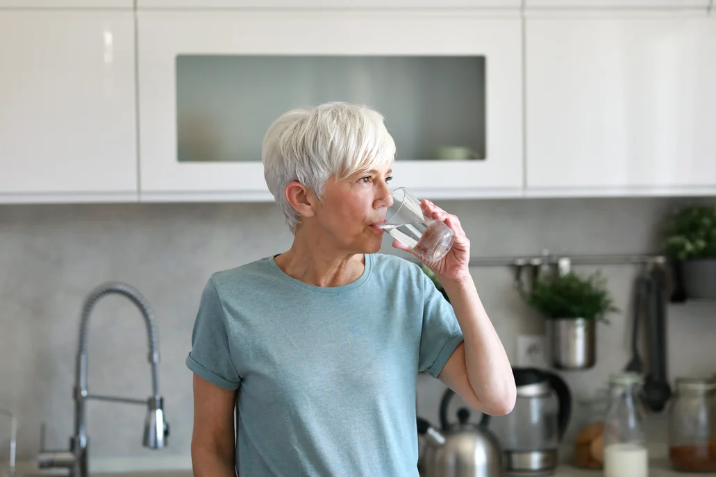 An older woman in a blue shirt drinking water out of a clear glass