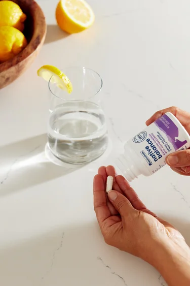 A hand pouring capsules of NativePath Native Balance from the container with a glass of water and lemon in the background.