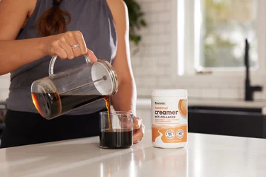 A woman pouring a mug of coffee with a container of NativePath Hazelnut Collagen Coffee Creamer next to it