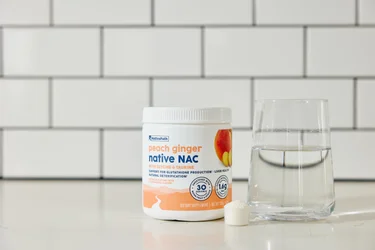 A frontal shot of a container of NativeNAC with a glass of water and a scoop next to it.