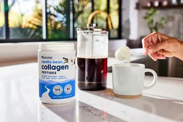 A hand pouring a scoop of NativePath Vanilla Bean collagen into a coffee mug with a French Press in the background