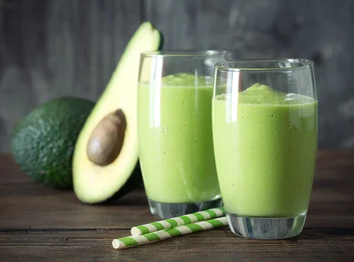 Two glasses of a green smoothie with avocado in the background