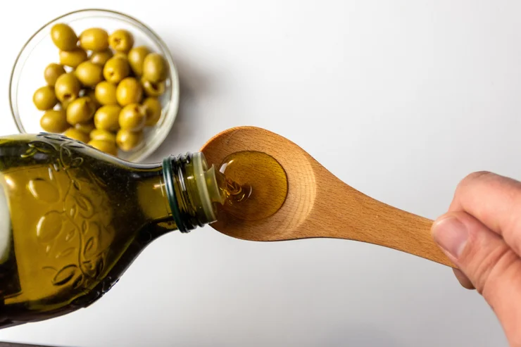 Olive oil being poured onto a wooden sppon with a bowl of olive in the background