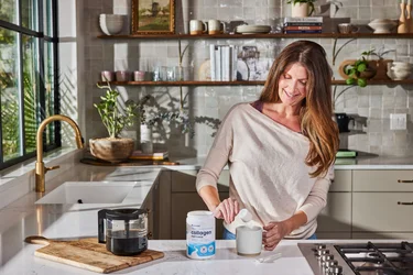 A woman in a kitchen pouring a scoop of NAtivePath Original Collagen Peptides into a coffee mug