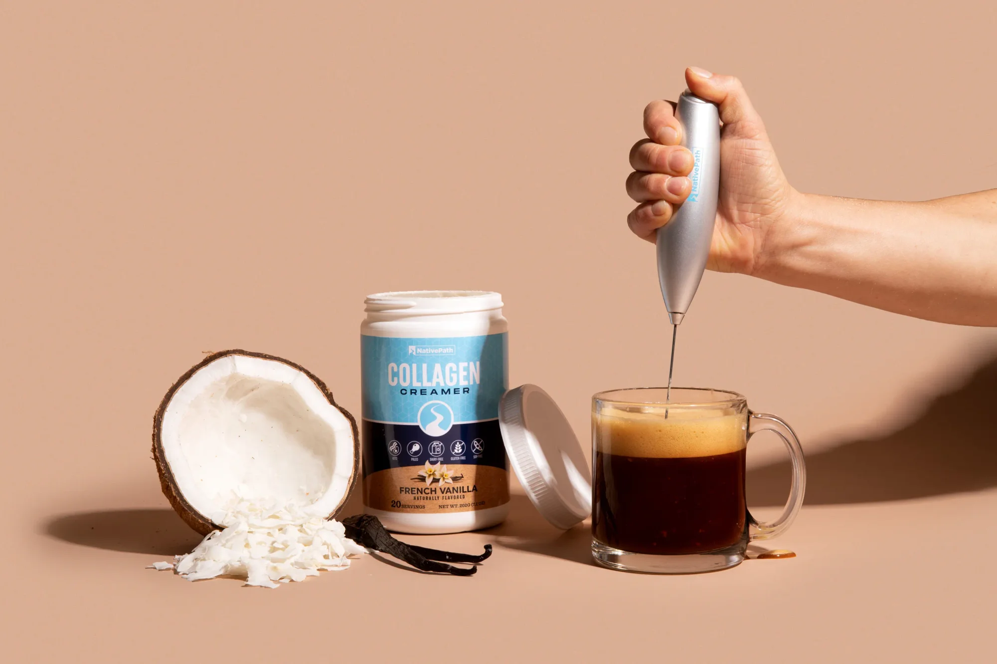 Hand frothing a scoop of NativePath Collagen Creamer in a clear cup of black coffee. Collagen creamer jar next to mug with vanilla bean and a halved coconut next to it.