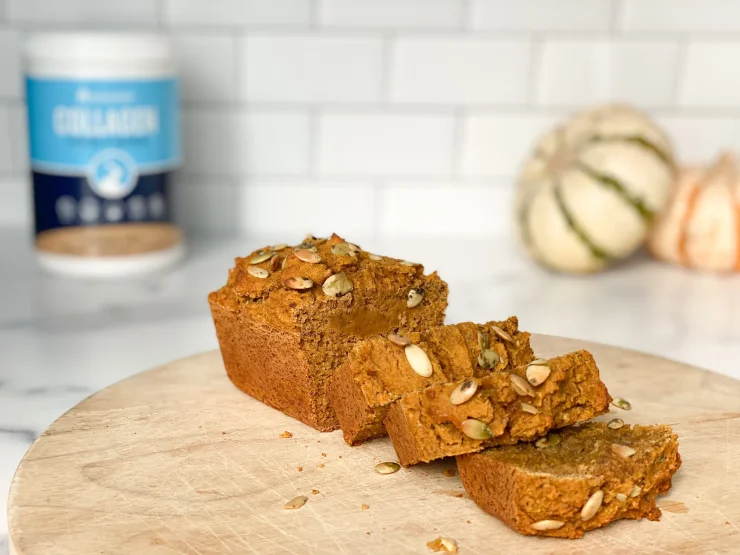 Paleo protein pumpkin bread with collagen on a wooden cutting board with small pumpkins and a collagen jar in the background.