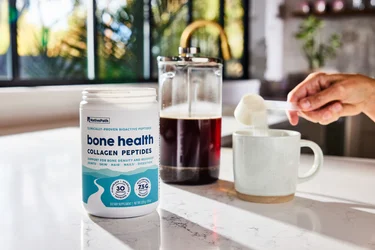 A hand pouring a scoop of NativePath Bone Health collagen into a coffee mug with a french press in the background