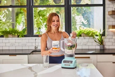 A woman pouring a scoop of NativePath MCT Powder into a blender full of fruits and veggies