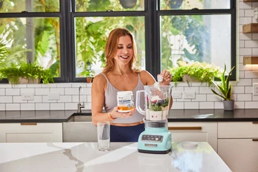 A woman pouring a scoop of NativePath MCT powder into a blender full of fruits and veggies