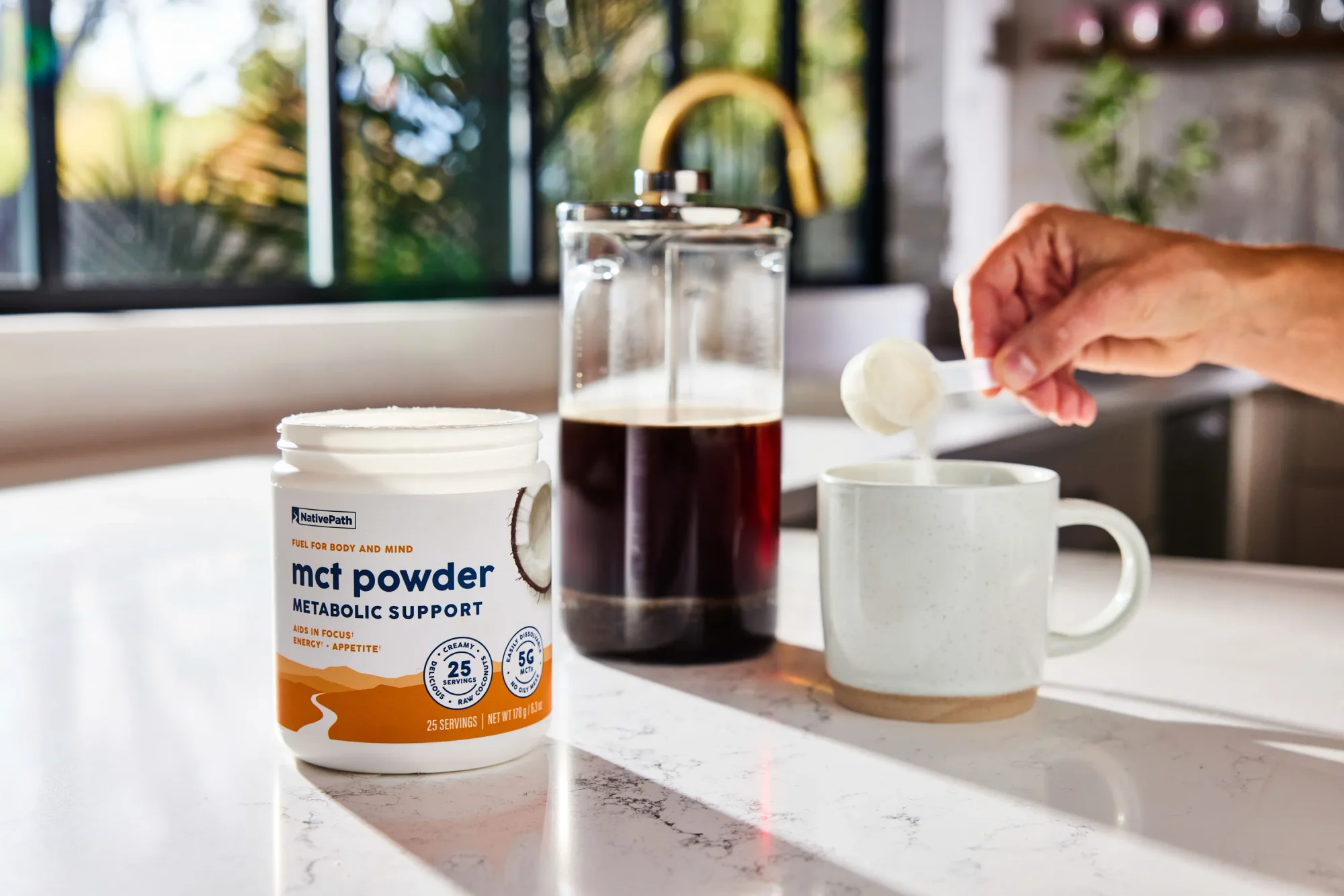 Hand pouring a scoop of NativePath Hazelnut Coffee Creamer into a cup of coffee. French press in the background.