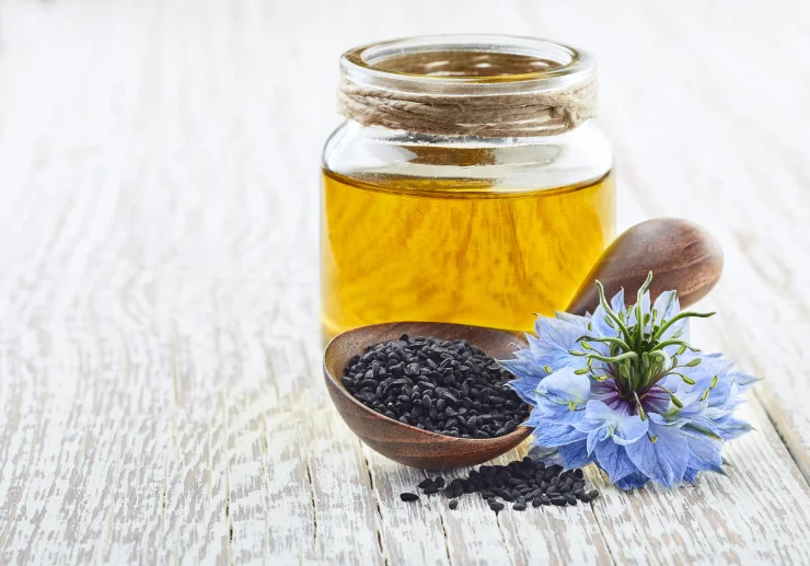 Clear jar of black seed oil, wooden spoon full of black seeds, and purple Nigella Sativa flower on white wooden board.