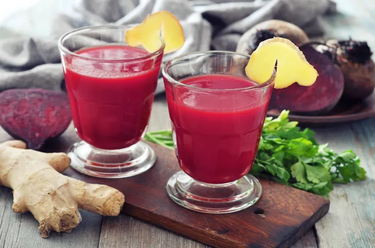 Two clear glasses of Ginger Beet Detox Juice on wooden cutting board. Ginger root and sliced beets in background