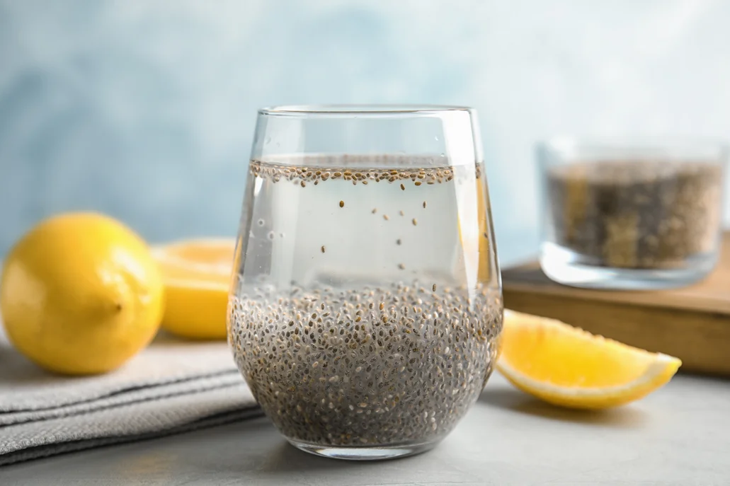 A glass of water with chia seeds soaking. A bowl of chia seeds and lemons in the background.