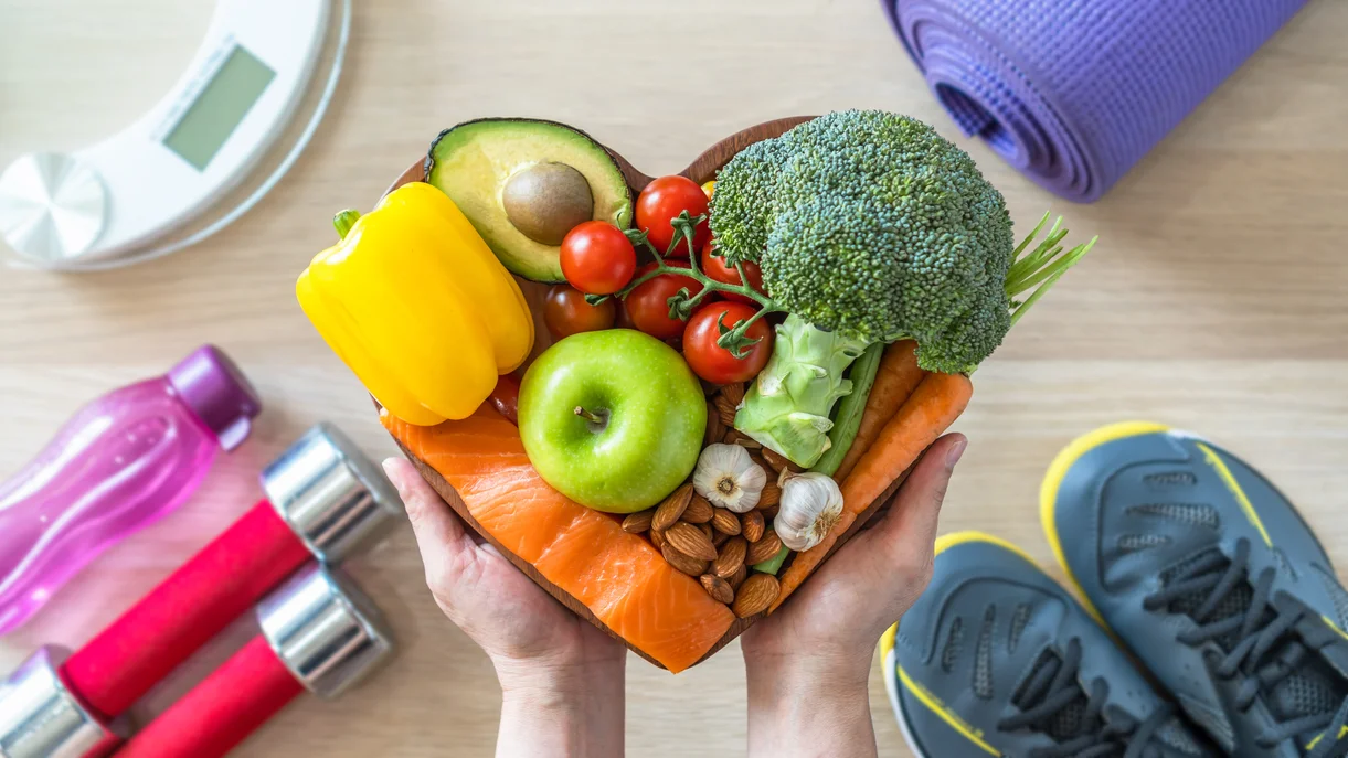 A person holding a heart shaped plate full of heart healthy food, with exercise equipment in the background
