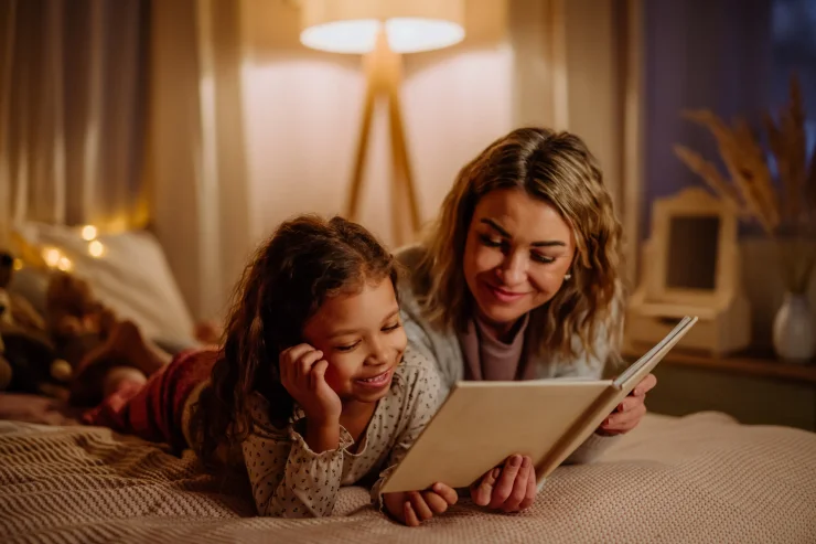 Happy mother with her little daughter lying on bed and reading a book in the evening at home.