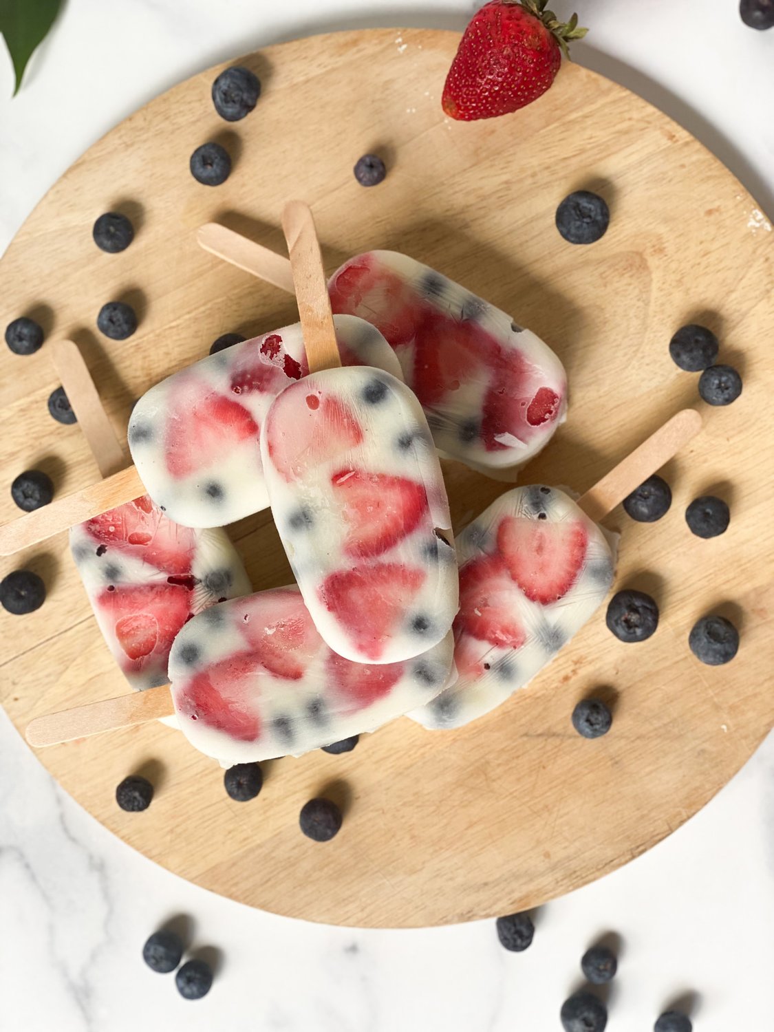 Berry yogurt pops stacked on a circular wooden board with blueberries scattered